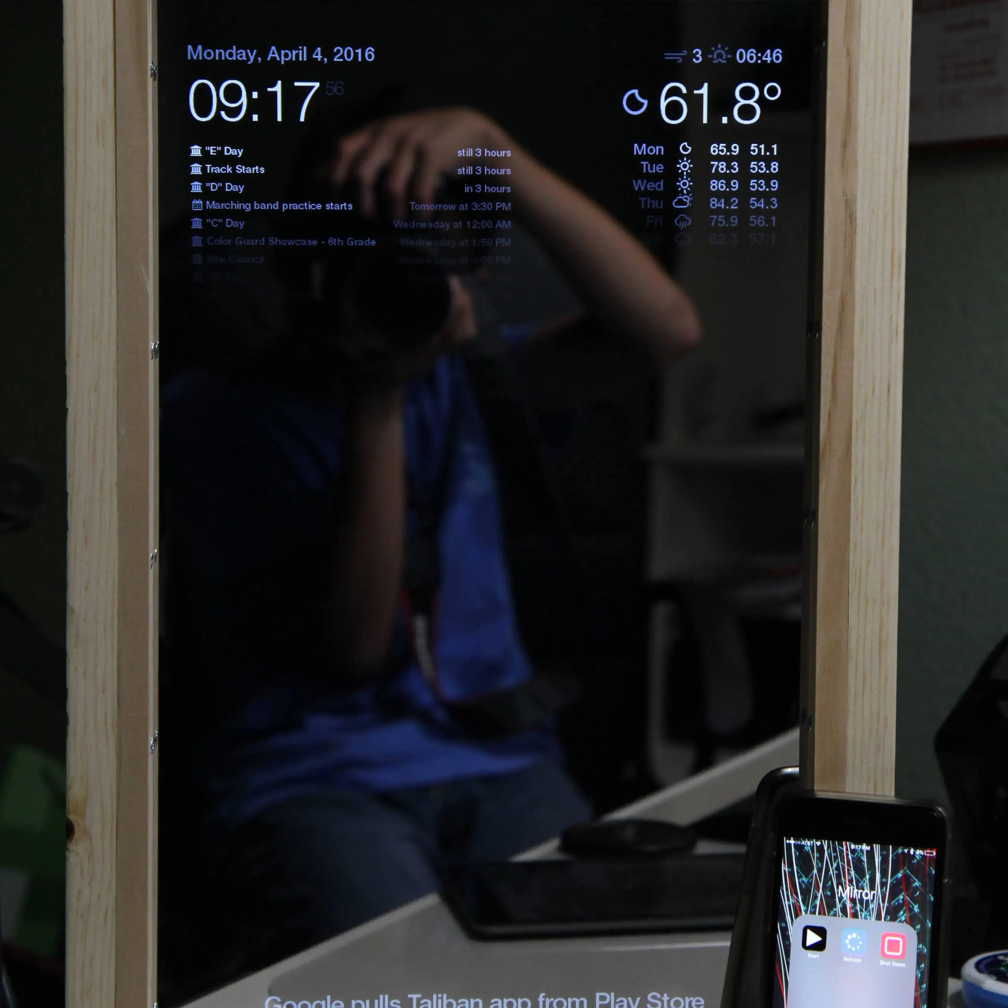 A shot of the front of the Magic Mirror. In the mirror's reflection, Kyle is seen holding his DSLR camera to take a picture of the Magic Mirror. The Mirror magically shows information 'through' the mirror: on the top, the time, date, weather, weekly forecast, calendar events are shown. News headlines are shown at the bottom of the mirror. Alongside the mirror is an iPhone 5. The iPhone shows a folder on the home screen, with the folder's name being 'Mirror'. The folder contains three shortcuts: 'Start', 'Restart', and 'Shut Down'. The start button icon is a white play icon on a black background. The restart button icon is a white circular pattern on a blue background. The shut down icon is a white, hollow square on a red background.